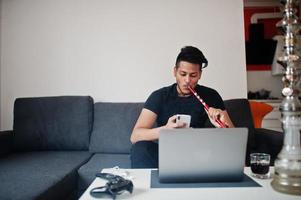 hombre indio guapo y de moda en negro sentado en la habitación, fumando narguile, mirando el teléfono con la computadora portátil. foto