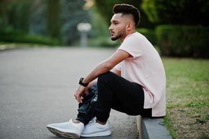 Stylish indian beard man at pink t-shirt. India model sitting at green garden of city. photo