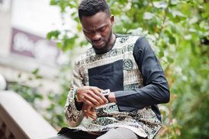 apuesto hombre afroamericano vestido con ropa tradicional en una ciudad moderna mirando sus relojes. foto