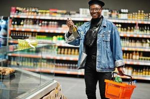 elegante hombre afroamericano casual con chaqueta de jeans y boina negra sosteniendo una canasta y mirando una botella de vino, comprando en el supermercado. foto