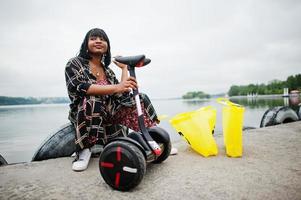 hermosa mujer afroamericana sentada cerca de segway o hoverboard. chica negra con símbolo de reciclaje de bolsas ecológicas de tela amarilla. foto