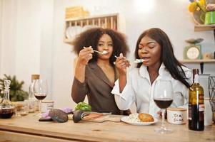 Two african american female friends women spending time at kitchen with wine. Black girlfriends  relaxing at home and eat salad. photo