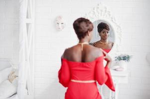 modelo afroamericano de moda en vestido de belleza rojo, mujer sexy posando vestido de noche y mirando al espejo. foto