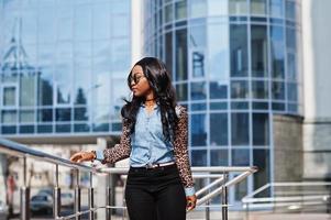 chica afroamericana hipster con gafas de sol, camisa de jeans con mangas de leopardo posando en la calle contra un edificio de oficinas moderno con ventanas azules. foto