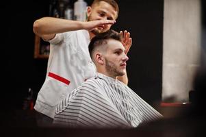 Young bearded man getting haircut by hairdresser while sitting in chair at barbershop. Barber soul. photo