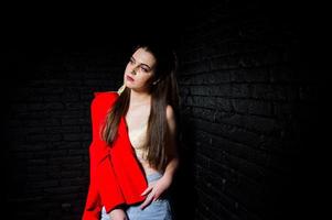 chica morena con estilo en chaqueta roja contra la pared de ladrillo negro de estudio. foto