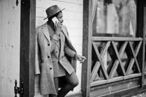 Stylish African American man model in gray coat, jacket tie and red hat posed against wooden cafe and speaking on mobile phone. Black and white photo. photo
