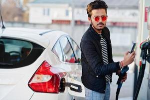 South asian man or indian male refueling his white car on gas station and pay by mobile phone. photo