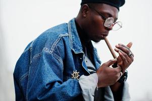 hombre afroamericano con chaqueta de jeans, boina y anteojos, enciende un cigarro. foto