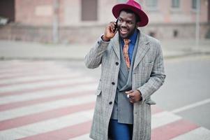 elegante modelo afroamericano con abrigo gris, corbata de chaqueta y sombrero rojo caminando en el cruce de peatones y hablando por teléfono móvil. foto