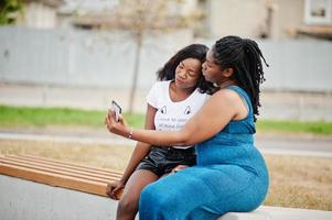 Two african american dark skinned friends female. One of them plus size model, second slim. Having fun and spending time together. Look at mobile phone. photo