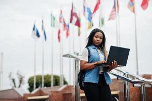 African student female posed with backpack and school items on yard of university, against flags of different countries. She hold laptop on hands. photo