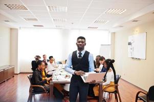 Face of handsome african business man, holding laptop on the background of business peoples multiracial team meeting, sitting in office table. photo