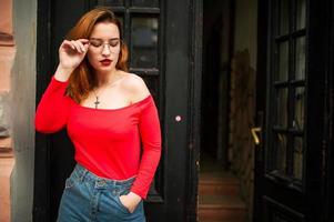 Attractive redhaired woman in eyeglasses, wear on red blouse and jeans skirt posing at street against old wooden door. photo