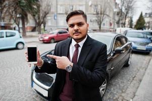 Stylish indian businessman in formal wear standing against black business car on street of city and show finger to mobile phone screen. photo