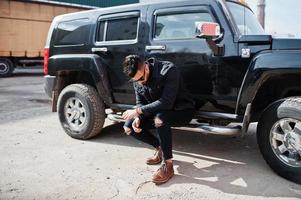 Fashion rich beard Arab man wear on black jeans jacket and sunglasses posed against big black suv car. Stylish, succesful and fashionable arabian model guy. photo