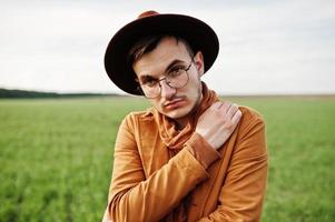 Stylish man in glasses, brown jacket and hat posed on green field. photo