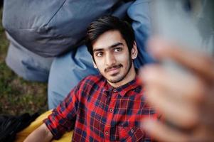 Young indian student man at checkered shirt and jeans sitting and relax at outdoor pillows and making selfie. photo