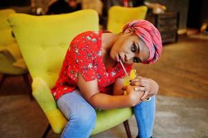 elegante mujer africana con camisa roja y sombrero posó en un café interior y bebiendo limonada de piña. foto