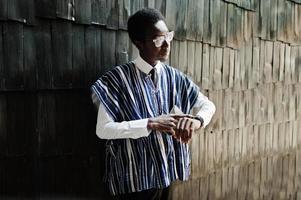 African businessman in traditional clothes and glasses with mobile phone at hand looking at his watches against wooden wall. photo