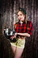 Young funny housewife in checkered shirt and yellow shorts pin up style with kitchen sieve on wooden background. photo