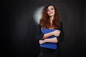 Brunette curly haired girl in black office jacket with skirt, on bra with folder of documentts at hands on studio against black background. Sexy businesswoman. photo