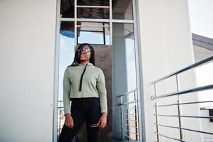 City portrait of positive young dark skinned female wearing green hoody and eyeglasses standing at balkony. photo