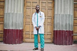 médico afroamericano masculino en bata de laboratorio con estetoscopio al aire libre contra la puerta de la clínica. foto