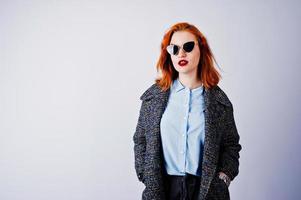 retrato de una fantástica chica pelirroja con camisa azul, abrigo gris posando con gafas de sol en el estudio. foto