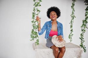 Attractive african american woman with afro hair wear on skirt and jeans jacket, posed at white room on swing. Fashionable black model. photo