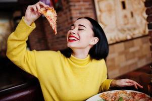 chica morena divertida en suéter amarillo comiendo pizza en el restaurante. foto