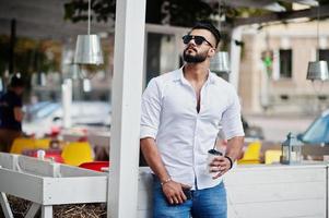 Stylish tall arabian man model in white shirt, jeans and sunglasses posed at street of city. Beard attractive arab guy with cup of coffee against outdoor cafe. photo