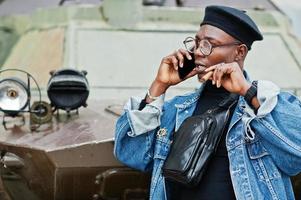 African american man in jeans jacket, beret and eyeglasses, with cigar posed against btr military armored vehicle, and speaking on mobile phone. photo