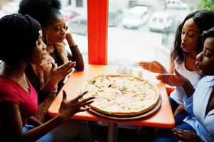 cuatro jóvenes africanas en un restaurante de colores brillantes oliendo pizza y divirtiéndose juntas. foto