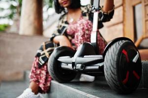 hermosa mujer afroamericana sentada cerca de segway o hoverboard. chica negra con scooter eléctrico autoequilibrado de doble rueda. foto