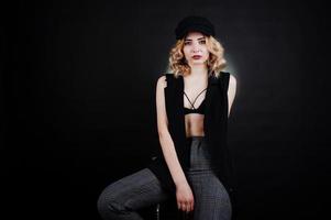 Studio portrait of blonde girl in black wear, bra and cap against dark background. photo