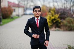 joven indio con anteojos, traje negro con corbata roja posado al aire libre. foto