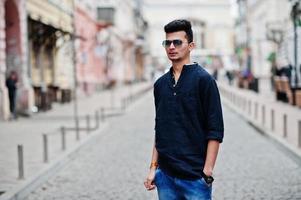 elegante modelo indio con ropa informal, camisa negra y gafas de sol posadas al aire libre en la calle de la india. foto