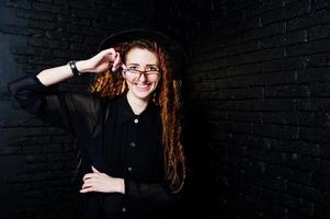 sesión de estudio de una chica de negro con rastas, anteojos y sombrero sobre fondo de ladrillo. foto