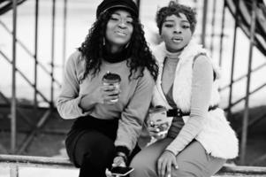 Two african american womans posed at winter day against snowy background with cups of coffee. photo