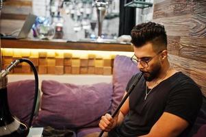 hombre árabe de barba elegante con gafas y camiseta negra fumando narguile en el bar interior. modelo árabe descansando. foto