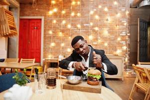 Respectable young african american man in black suit sitting in restaurant with tasty double burger and soda drink. Divide half by knife. photo