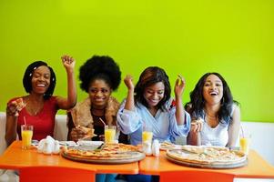 Four young african girls in bright colored restaurant eating pizza and having fun together. photo