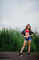 Sexy curly model girl in red top, jeans denim shorts, leather jacket and sneakers posed in the reed. photo