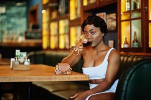 African american woman, retro hairstyle in white dress at restaurant with glass of wine. photo