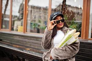 Fashionable african american woman in black cap, sunglasses and fur with two baguette rolls at hands. photo