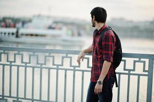 joven estudiante indio con camisa a cuadros y jeans con mochila posada en la ciudad nocturna contra el lago y el barco. foto