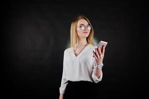 Studio portrait of blonde businesswoman in glasses, white blouse and black skirt looking at  mobile phone against dark background. Successful woman and stylish girl concept. photo