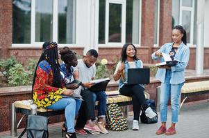 Group of five african college students spending time together on campus at university yard. Black afro friends studying at bench with school items, laptops notebooks. photo