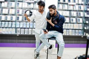 Two indians mans customer buyer at mobile phone store with earphones and wireless speaker listening music. South asian peoples and technologies concept. Cellphone shop. photo